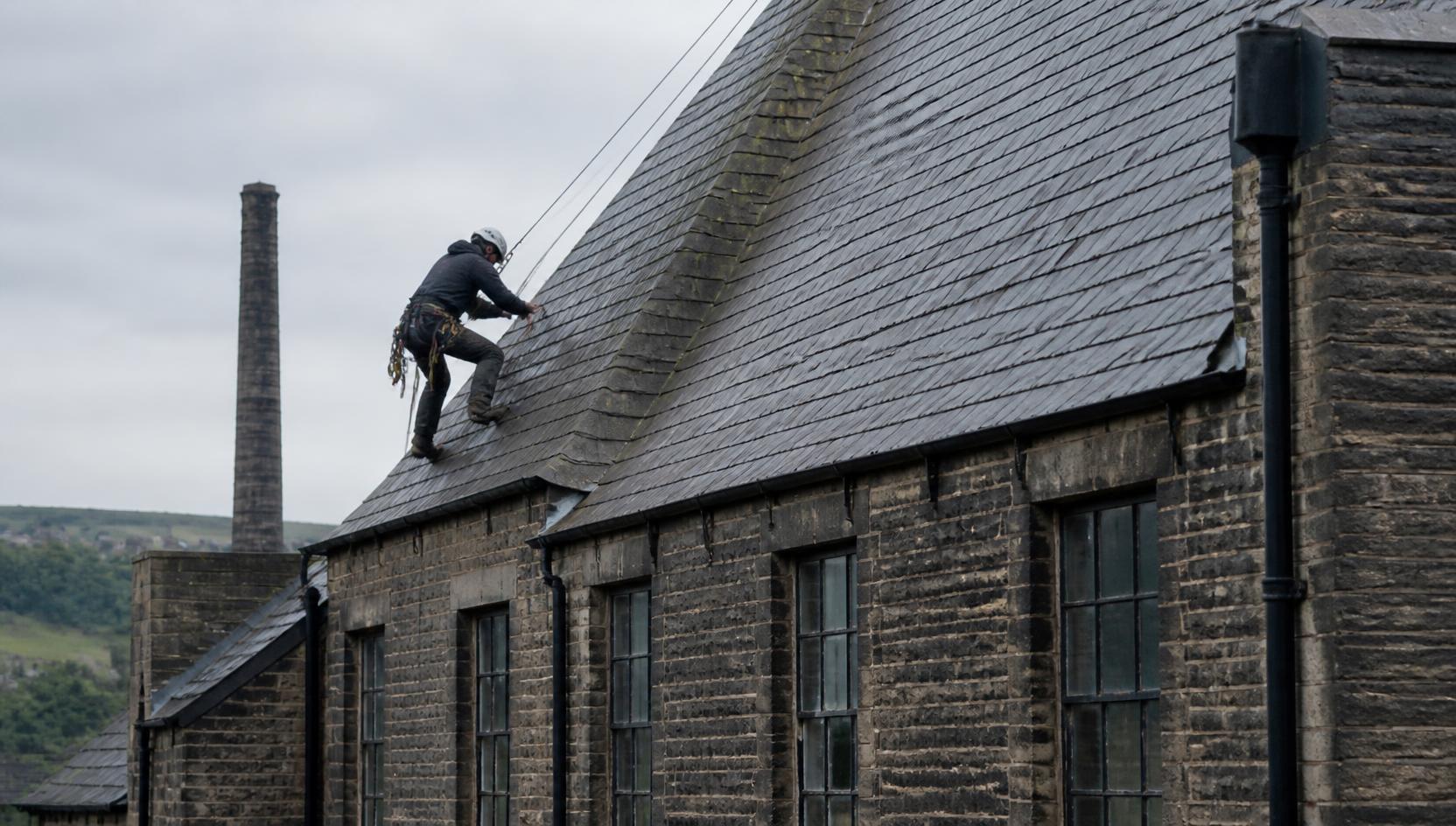 Rope access technician working on the steep slate roof of an older stone mill-style building in Bolton